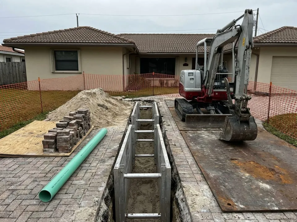 Mini excavator and shored trench cut through a paver driveway during sewer line replacement at a Florida house